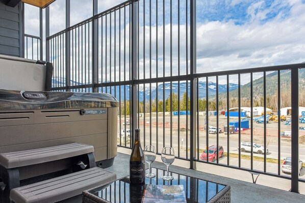 A balcony with a grill, a bottle of wine, and glasses, overlooking a parking area and mountains. - Revy Mountain Dream (Revelstoke)