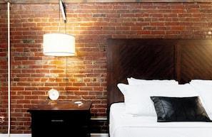 A hotel room with a brick wall, a wooden headboard, a bedside table with a clock, and a bed with white bedding and a black pillow. - *New* Wings Over Nulu | Luxury 2BR Industrial Loft (Louisville)