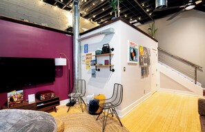 A room with a flat-screen TV, a chair, a small table with a radio, and a wall with posters and a shelf. - *New* Wings Over Nulu | Luxury 2BR Industrial Loft (Louisville)