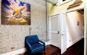 A room with a brick wall, a blue chair, a white door, and a painting of an airplane. - *New* Wings Over Nulu | Luxury 2BR Industrial Loft (Louisville)