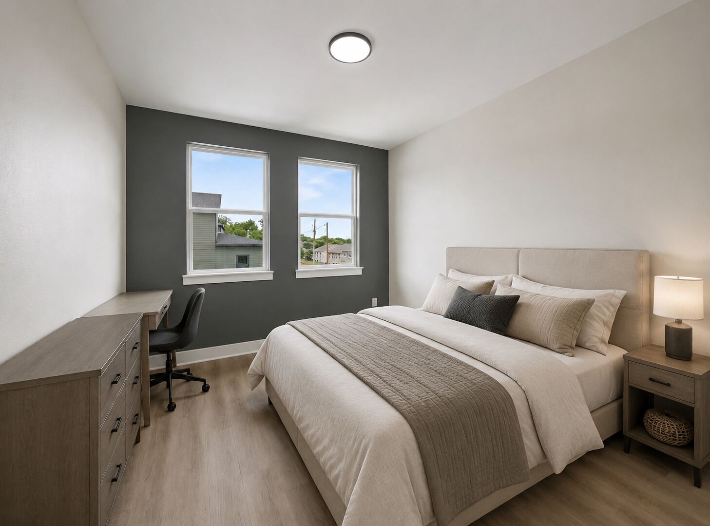 Ein Schlafzimmer mit Bett, Nachttisch, Lampe, Schreibtisch, Stuhl und zwei Fenstern.