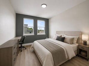 Ein Schlafzimmer mit Bett, Nachttisch, Lampe, Schreibtisch, Stuhl und zwei Fenstern.