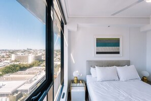Ein modernes Hotelzimmer mit großem Fenster, einem Bett mit weißer Bettwäsche, einem Nachttisch mit Lampe und einem Bild an der Wand.