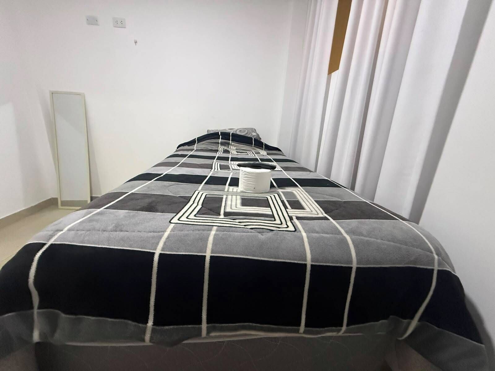 Habitación de hotel estrecha con una cama centrada, una mesita de noche y una cortina a un lado.