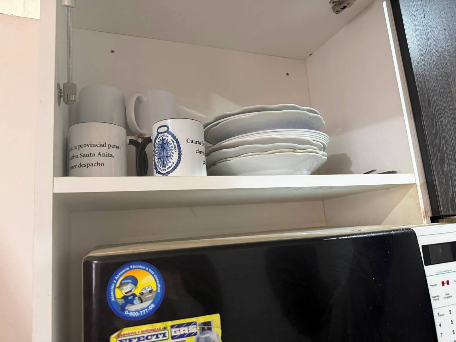 Uma cozinha com geladeira, uma prateleira com pratos e uma caneca, e um armário fixado na parede.