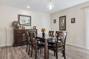 A dining area with a wooden table and chairs, a mirror, and framed artwork on the walls. - Rolling Hills Haven | 3BR | Large Screened Patio | Fenced Yard | Sun City | Sleeps 6 (Sun City)