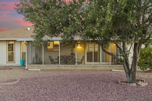 A house with a covered patio, a table and chairs, and a tree in the foreground. - Rolling Hills Haven | 3BR | Large Screened Patio | Fenced Yard | Sun City | Sleeps 6 (Sun City)