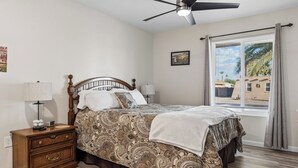 Ein Schlafzimmer mit Bett, Nachttischen, Deckenventilator, Fenster mit Jalousien und einem Wandbild.
