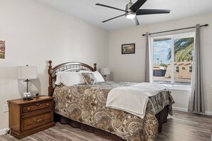 A bedroom with a bed, nightstands, a ceiling fan, a window with blinds, and a wall-mounted picture. - Rolling Hills Haven | 3BR | Large Screened Patio | Fenced Yard | Sun City | Sleeps 6 (Sun City)
