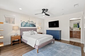 A bedroom with a bed, bedside tables, a ceiling fan, artwork, a TV, and a view into the kitchen.