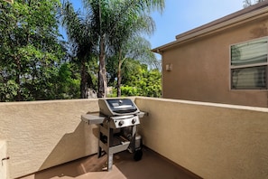 Una zona de parrilla al aire libre con una parrilla de gas portátil, un cubo de basura y una ventana con vistas a un jardín con árboles.