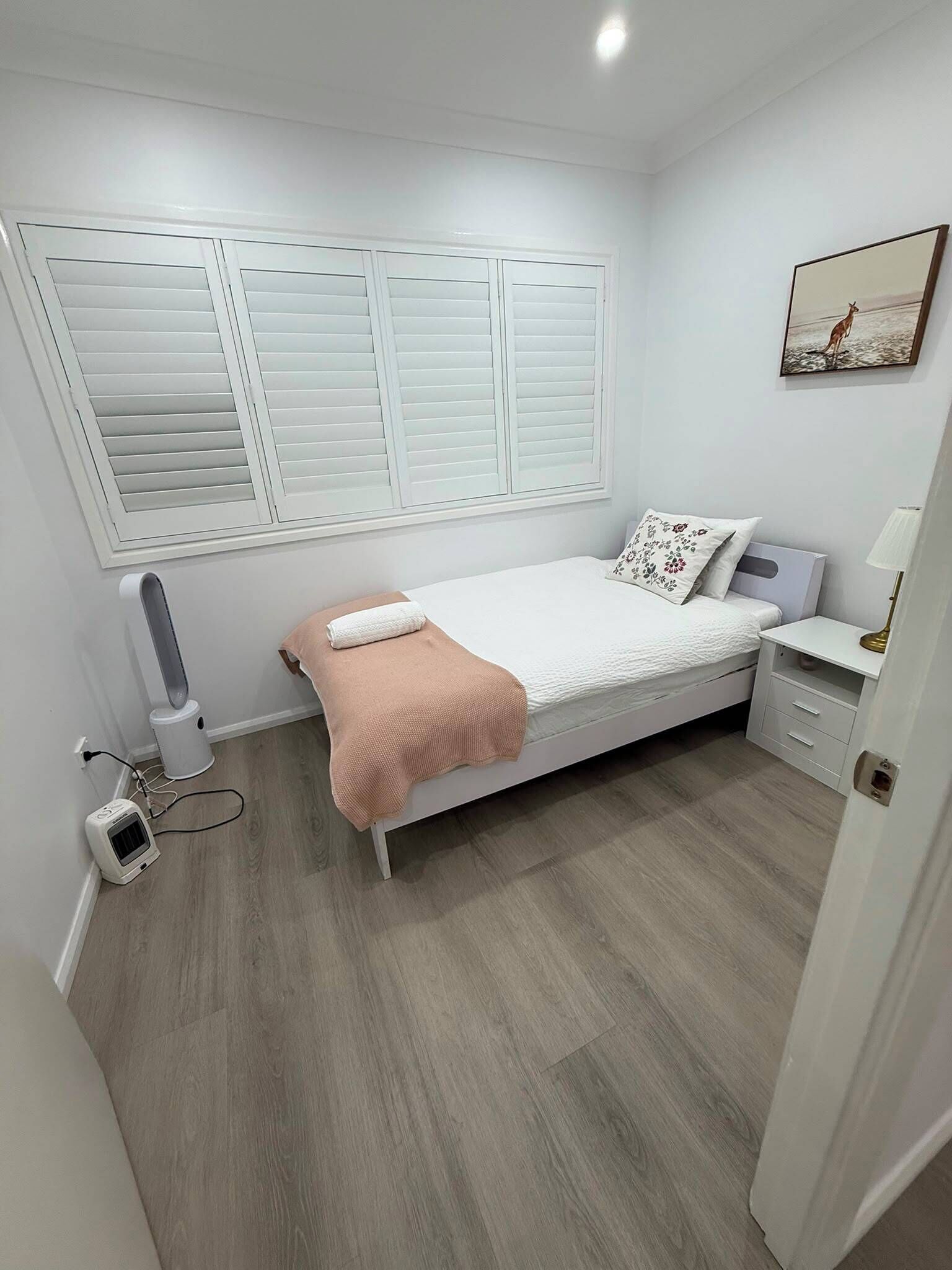 A bedroom with a bed, bedside table, lamp, heater, and a window with shutters.