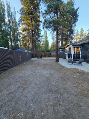 A fenced outdoor area with a gravel path, a wooden deck with chairs, and a cabin-like structure with a window. - Cozy Big Bear Cabin _Game Room-Pet Friendly- Near Lake-Sleeps 8 (Big Bear)