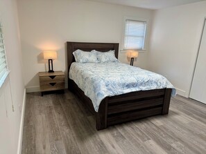 Un dormitorio con cama de madera, dos mesitas de noche con lámparas, una ventana con persianas y suelo de madera.