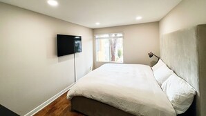 Ein Schlafzimmer mit einem Bett, einem an der Wand montierten Fernseher, einem Fenster mit Jalousien und einer Lampe auf dem Nachttisch.