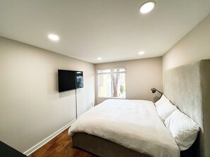 Een slaapkamer met een bed, een aan de muur bevestigde televisie, een raam met zonwering en een lamp op de nachtkastje.