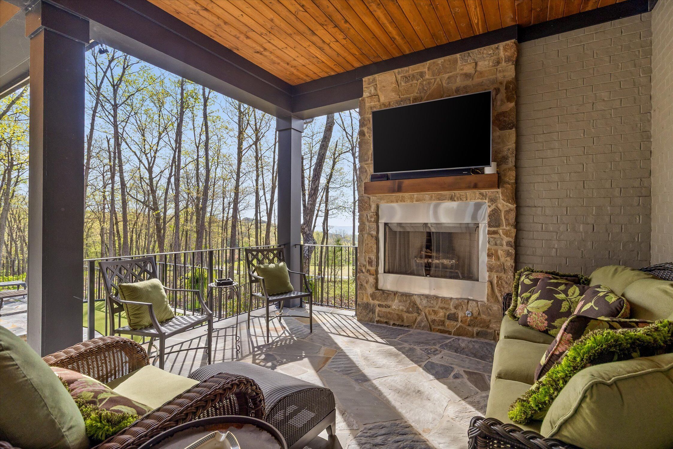 A covered outdoor seating area with a stone fireplace, a flat-screen TV mounted on the wall, and a view of the surrounding trees.