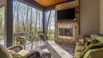 A covered outdoor seating area with a stone fireplace, a flat-screen TV mounted on the wall, and a view of the surrounding trees.