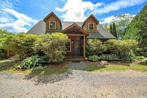 A two-story log cabin with a symmetrical facade, a gravel driveway, and a well-maintained garden with various shrubs and a bench. - Scenic Mountain Retreat Just Minutes from Boone (Boone)