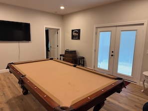 A pool table in a room with a television mounted on the wall, a door with a glass panel, and a small table with a barrel.