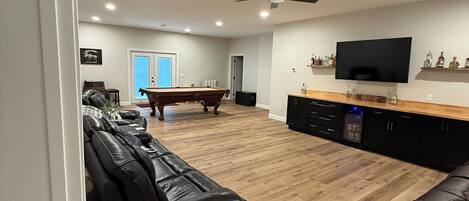 Amplia sala de estar con mesa de billar, sofás de cuero, ventilador de techo y televisor de pantalla plana montado en la pared.