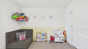 A cozy room with a grey sofa, a small bookshelf, a toy kitchen set, and a wall-mounted macrame decoration. - Owl's Head Haven 235-4 Bed House-Unfurnished (Freeport)
