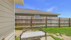 A patio area with a wicker chair and a small table, a wooden fence, and a house with a window. - Owl's Head Haven 235-4 Bed House-Unfurnished (Freeport)