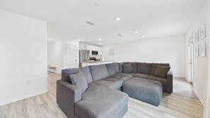 A modern living room with a sectional sofa, a kitchen in the background, and a hallway leading to a door. - Owl's Head Haven 235-4 Bed House-Unfurnished (Freeport)