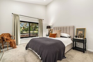Ein Schlafzimmer mit einem großen Bett, einem Nachttisch, einem Stuhl und Blick auf einen Garten durch ein Fenster.