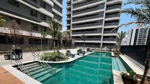 A swimming pool with a wooden deck, surrounded by palm trees and lounge chairs. - Apartamento com varanda - Pinheiros (São Paulo)