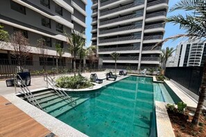 A swimming pool with a wooden deck, surrounded by palm trees and lounge chairs. - Apartamento com varanda - Pinheiros (São Paulo)