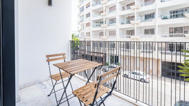 A balcony with a wooden table and chairs, a metal railing, and a view of a multi-story building with balconies. - Palmier • 1BR avec balcon & parking (Casablanca)