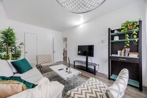 Ein modernes Wohnzimmer mit Sofa, Couchtisch und Fernseher. Pflanzen und ein Bücherregal mit Büchern sind ebenfalls vorhanden.