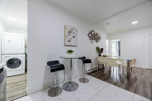 Uma cozinha moderna com máquina de lavar e secar embutidas, uma mesa de vidro redonda com dois banquetes e uma área de refeições com mesa e cadeiras de vidro.