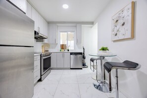 Ett modernt kök med ett kylskåp, en ugn och en diskmaskin i rostfritt stål, ett glasbord med två barstolar och en tavla med blommotiv som hänger på väggen.