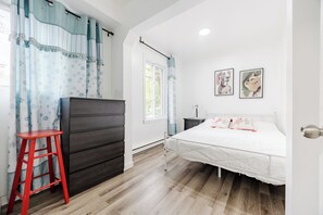 Quarto com cama, mesa de cabeceira, um banquinho vermelho, janela com cortinas e arte nas paredes.