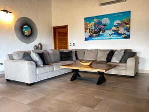 A living room with a grey sectional sofa, a wooden coffee table, and a colorful ocean-themed painting on the wall. - Sunset Villa Macaw, 2 King Suites, Pool & Ocean View (Manuel Antonio)
