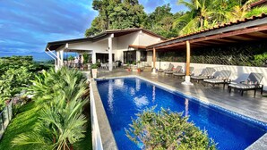 Et moderne poolområde med et klart blåt pool, liggestole og en overdækket terrasse.