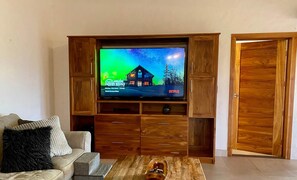 A wooden entertainment unit with a television displaying a show, a sofa with a throw pillow, and a coffee table with a decorative item. - Sunset Villa Macaw, 2 King Suites, Pool & Ocean View (Manuel Antonio)