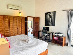 A bedroom with a bed, a desk, a chair, an air conditioner, a wall-mounted artwork, and a window with curtains. - Sunset Villa Macaw, 2 King Suites, Pool & Ocean View (Manuel Antonio)