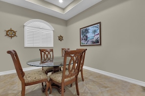 A dining area with a round glass table, wooden chairs, a window with shutters, a wall-mounted compass, and a framed picture. - Private Pool Villa | Players Club & Spa | Golf Views (Naples)