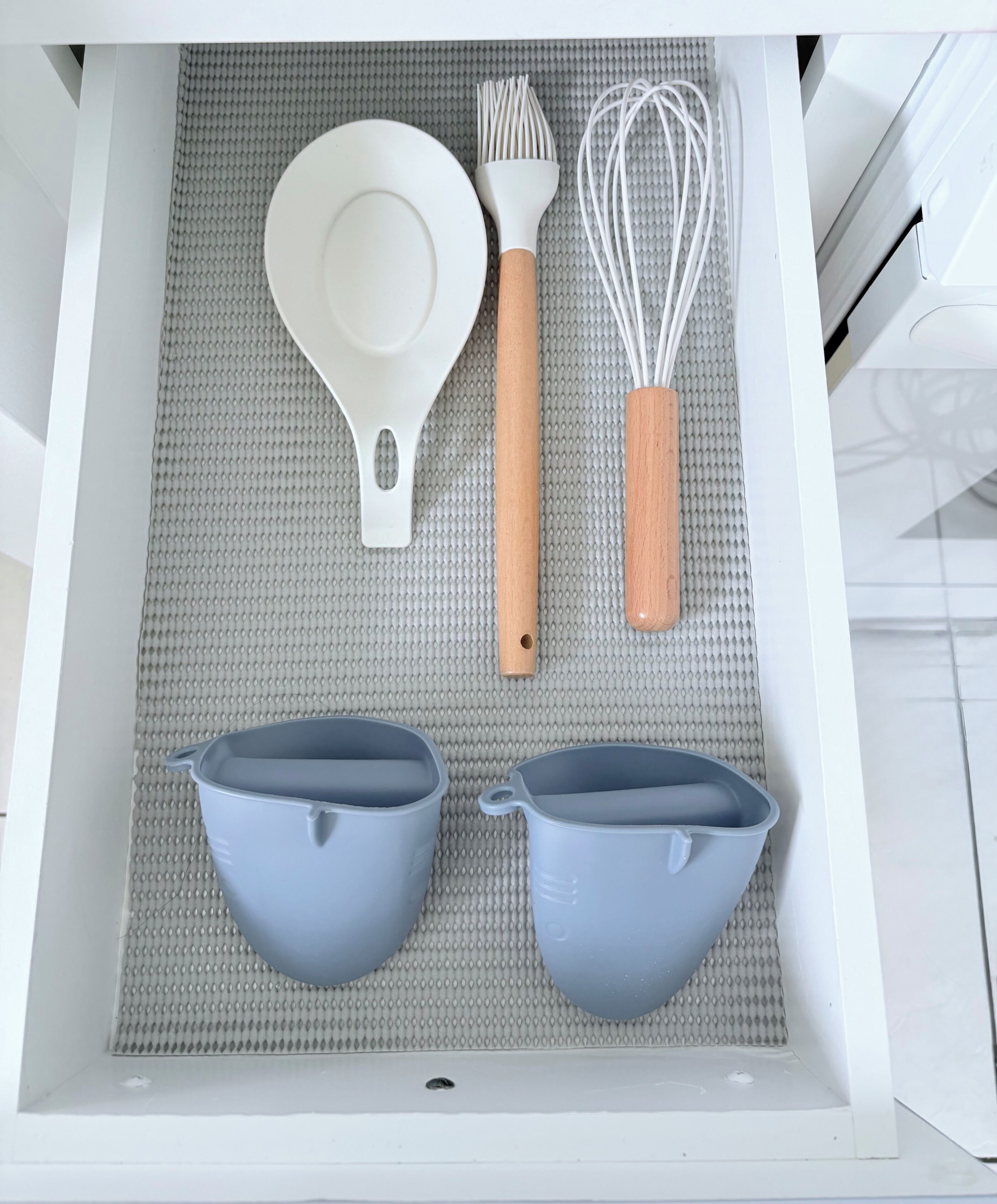 Un cajón de cocina con interior blanco, que contiene una espátula blanca, un batidor con mango de madera y dos tazas medidoras grises.