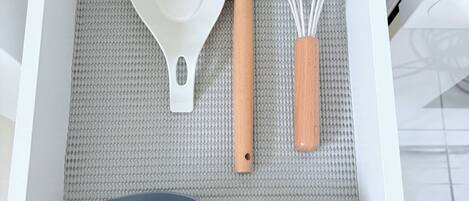 Un cajón de cocina con interior blanco, que contiene una espátula blanca, un batidor con mango de madera y dos tazas medidoras grises.