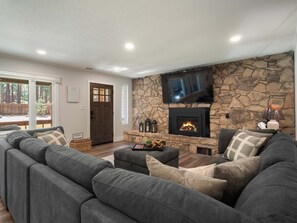 A living room with a stone wall, a fireplace, a flat-screen TV, and a large grey sofa. - Gabriel Getaway | Mountain Comfort in Skyland, NV (Zephyr Cove)