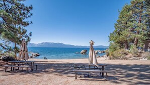 En strand med picknickbord och parasoller, en klar himmel och utsikt över havet och bergen.
