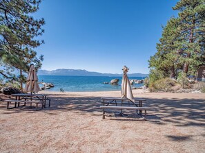 A beach with picnic tables and umbrellas, a clear sky, and a view of the sea and mountains. - Gabriel Getaway | Mountain Comfort in Skyland, NV (Zephyr Cove)