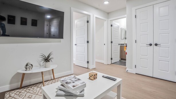 Una sala moderna con un televisor de pantalla plana, una pequeña mesa redonda con una planta, una mesa de centro con libros y un dado, y un pasillo que da a un dormitorio.