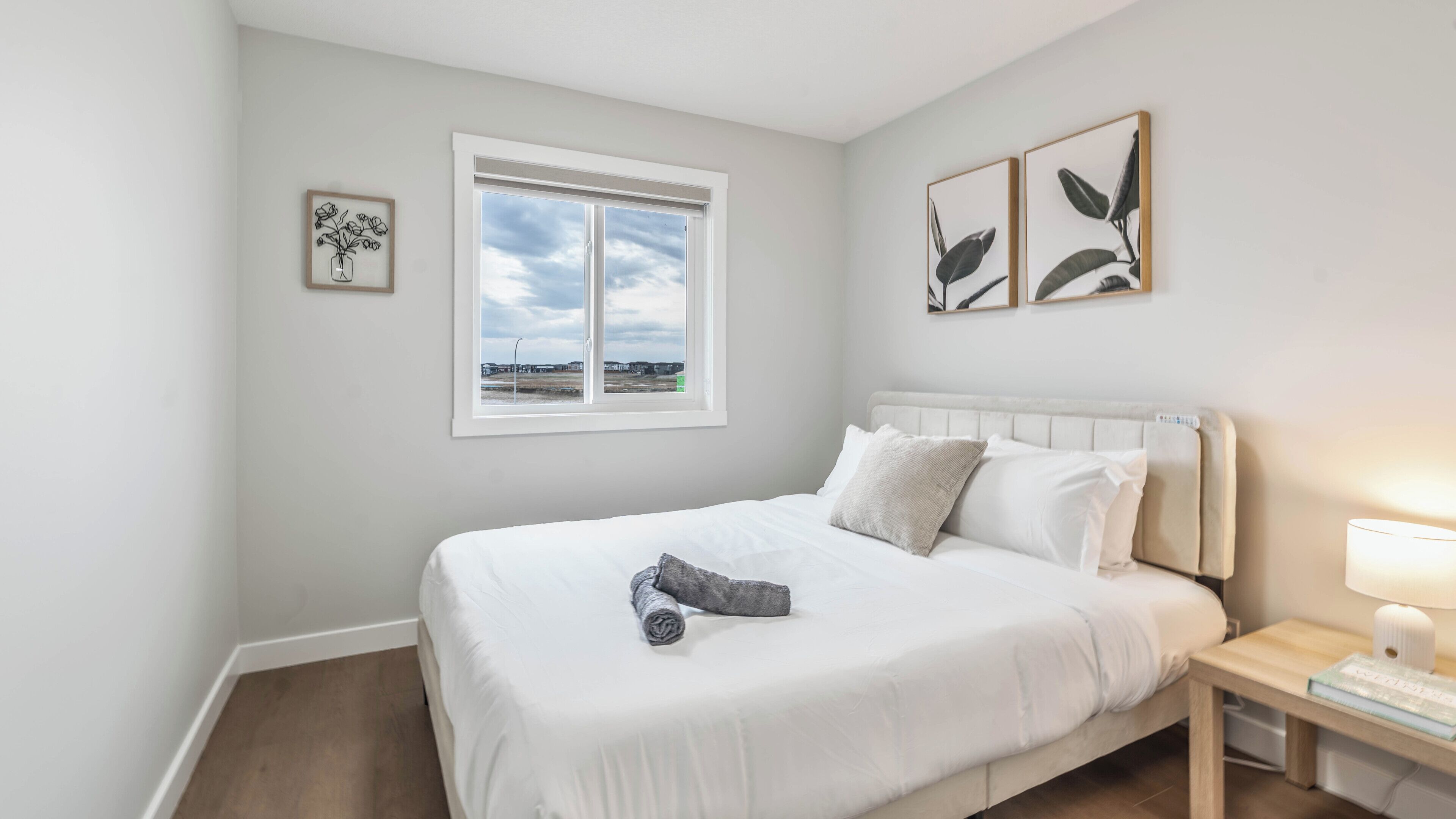 A bedroom with a bed, a nightstand, a window with a view, and two wall-mounted pictures.