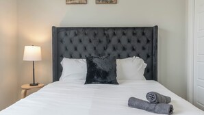 Una cama bien hecha con un cabecero capitonado, almohadas blancas, un cojín oscuro y dos toallas enrolladas.