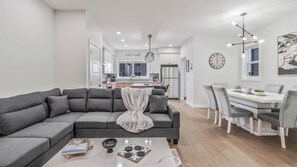 Ein modernes Wohnzimmer mit einem grauen Ecksofa, einem Essbereich mit weißem Tisch und grauen Stühlen sowie einer Küche im Hintergrund.